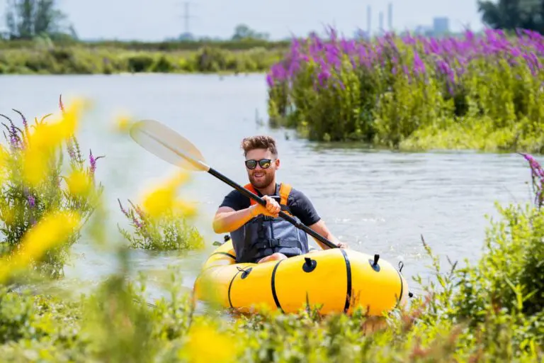 man in yellow packraft paddling through a river with purple flowers